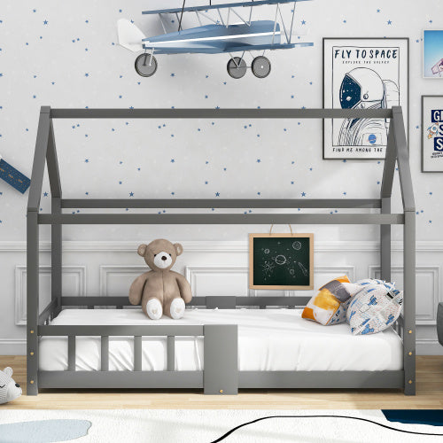 
Das Bild zeigt ein Kinderbett in Form eines Hauses, das in einem thematisch dekorierten Kinderzimmer steht. Das Bett ist grau und enthält eine Matratze mit Kissen. Ein brauner Teddybär sitzt auf dem Bett. An der Wand hinter dem Bett hängen Weltraum-Poster und ein Modellflugzeug. Eine kleine Tafel ist ebenfalls am Bett angebracht.