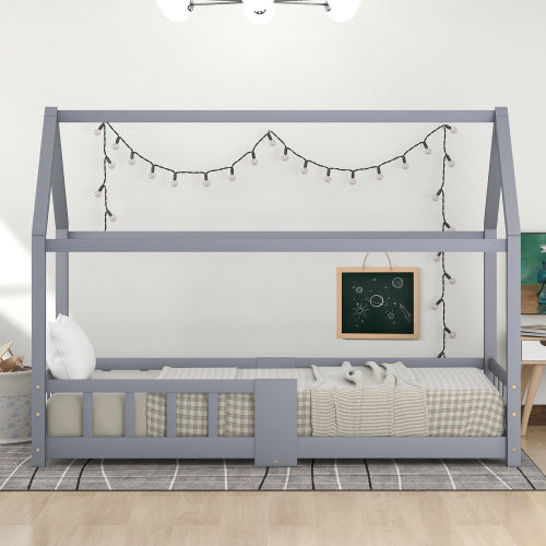 Das Bild zeigt ein Kinderbett in Form eines Hauses, das in einem schlicht eingerichteten Zimmer steht. Das Bett ist grau und mit einer Matratze, Kissen und einer karierten Decke ausgestattet. Über dem Bett sind Lichterketten aufgehängt. Eine kleine Tafel ist an der Bettseite angebracht. Der Boden ist mit einem Teppich bedeckt.