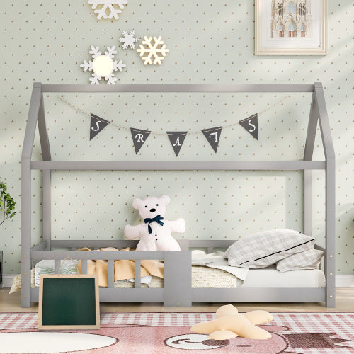 Das Bild zeigt ein Kinderbett in Form eines Hauses, das in einem gemütlichen Kinderzimmer steht. Das Bett ist grau und dekoriert mit einer Wimpelkette. Im Bett sitzt ein weißer Teddybär, und es gibt verschiedene Kissen und Decken. Der Hintergrund ist mit gepunkteter Tapete und Schneeflocken-Deko verziert. Auf dem Boden liegt ein Teppich mit einem Schachbrettmuster.